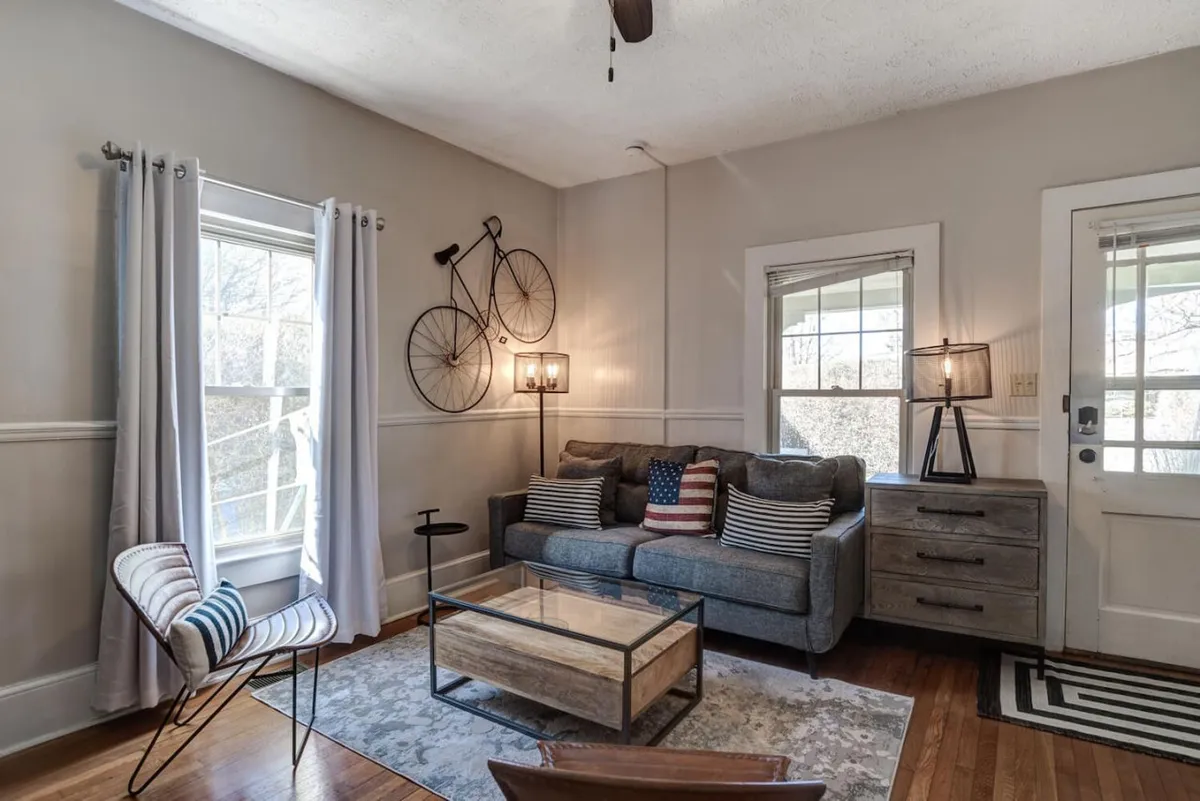 This image features a cozy living space with a plush sofa, bicycle wall art, a coffee table, a lamp, and a window streaming in afternoon light. The room feels designed for relaxing, reading, or enjoying a coffee, echoing the welcome guests receive upon entering.