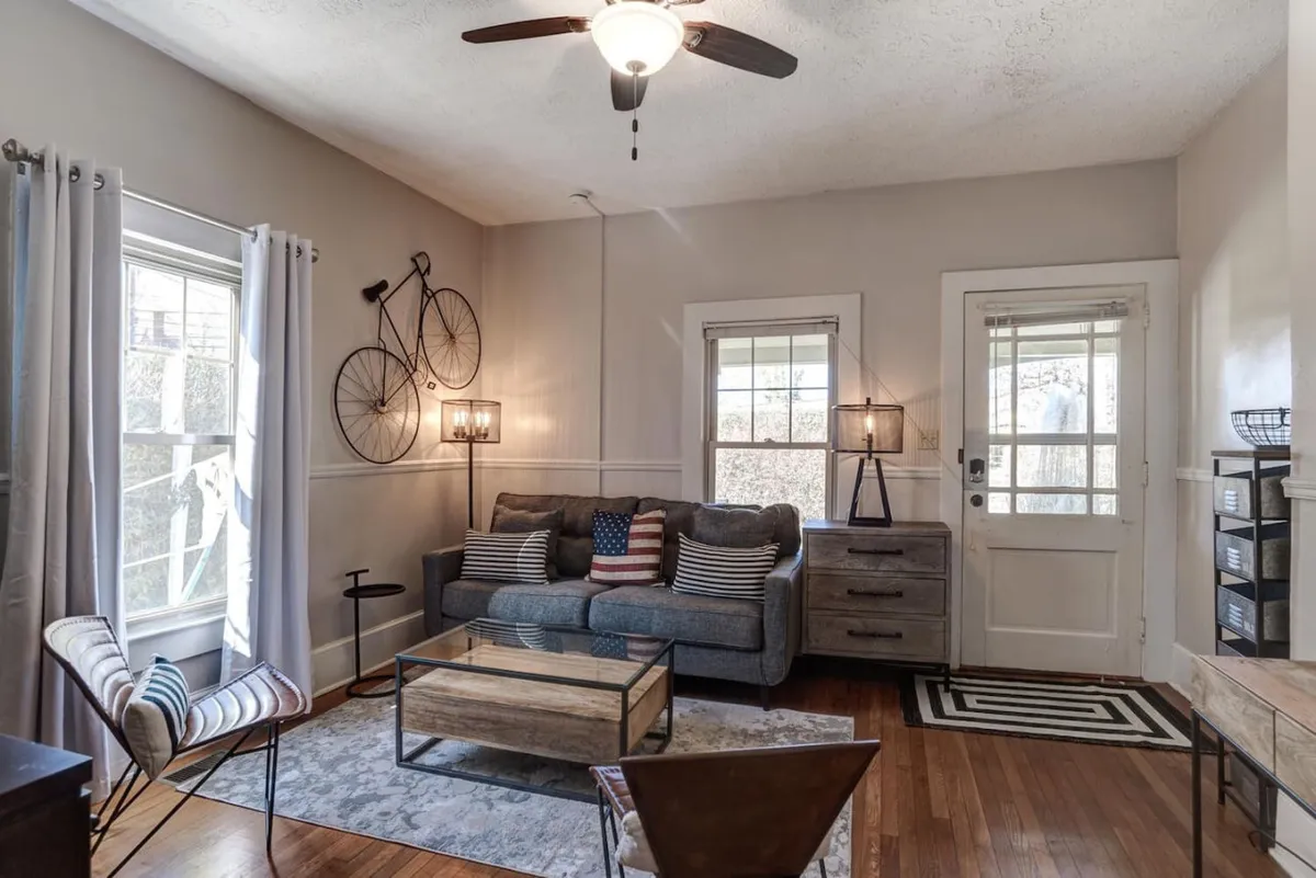 This image showcases a comfortable entry area with a sofa adorned with cushions, a sturdy wooden coffee table, bicycle-themed wall art, a floor lamp, and windows letting in natural light. The overall atmosphere is inviting and homey, suggesting a welcoming retreat immediately inside the private entrance.