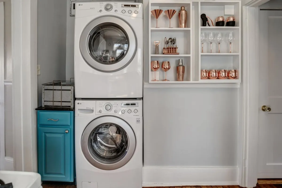 This image showcases a bright, cheerful interior space with a stacked washer and dryer alongside a stylish copper-accented shelving unit and a colorful cabinet. The room feels welcoming and practical, suggesting convenience for guests during longer stays.