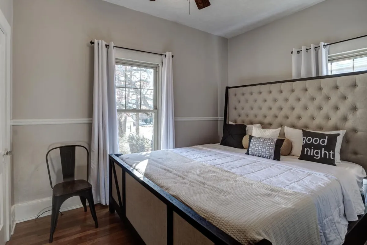 A cozy bedroom featuring a bed with a tufted headboard bathed in natural light from a nearby window. Decorative pillows and soft linens complete the restful ambiance, inviting relaxation and quiet moments like reading or unwinding after a busy day.