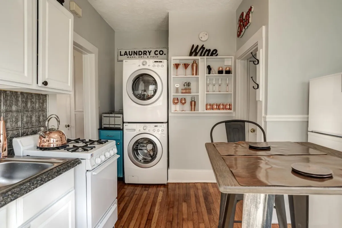 This interior scene presents a multifunctional space where a washing machine, dining table, decorative shelving, and kitchen appliances come together harmoniously. The area feels inviting yet practical, with natural light brightening the furnishings. Subtle decorative accents and functional furniture evoke a sense of historic character and flexible living—an ideal backdrop for sipping coffee, reading, or working.