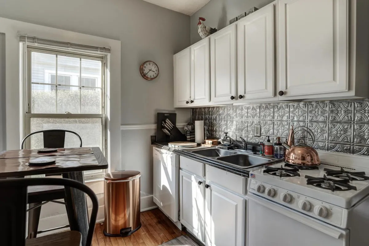 This image shows a charming, well-equipped kitchen featuring a kitchen sink, stove, and a gleaming copper kettle. A small dining table is also visible, evoking a warm and inviting mood ideal for both cooking and gathering for intimate meals. The overall setting is cozy and functional, highlighting the opportunity to prepare meals and enjoy them in a comfortable nook.