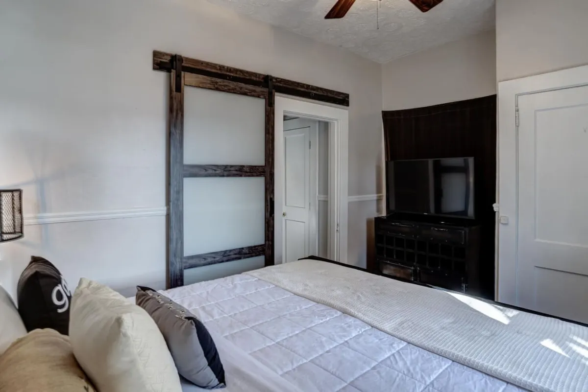 A calm, inviting bedroom scene with a neatly made bed dressed in clean bedding, complemented by a wooden sliding door and a sleek black entertainment unit. Decorative pillows add a touch of elegance and warmth, reinforcing the room's serene character.